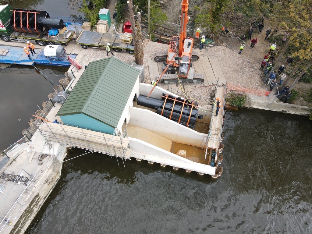 Drone vie of a turbine being lifted into its channel by a crane. Second turbine still on a trailer in the background.